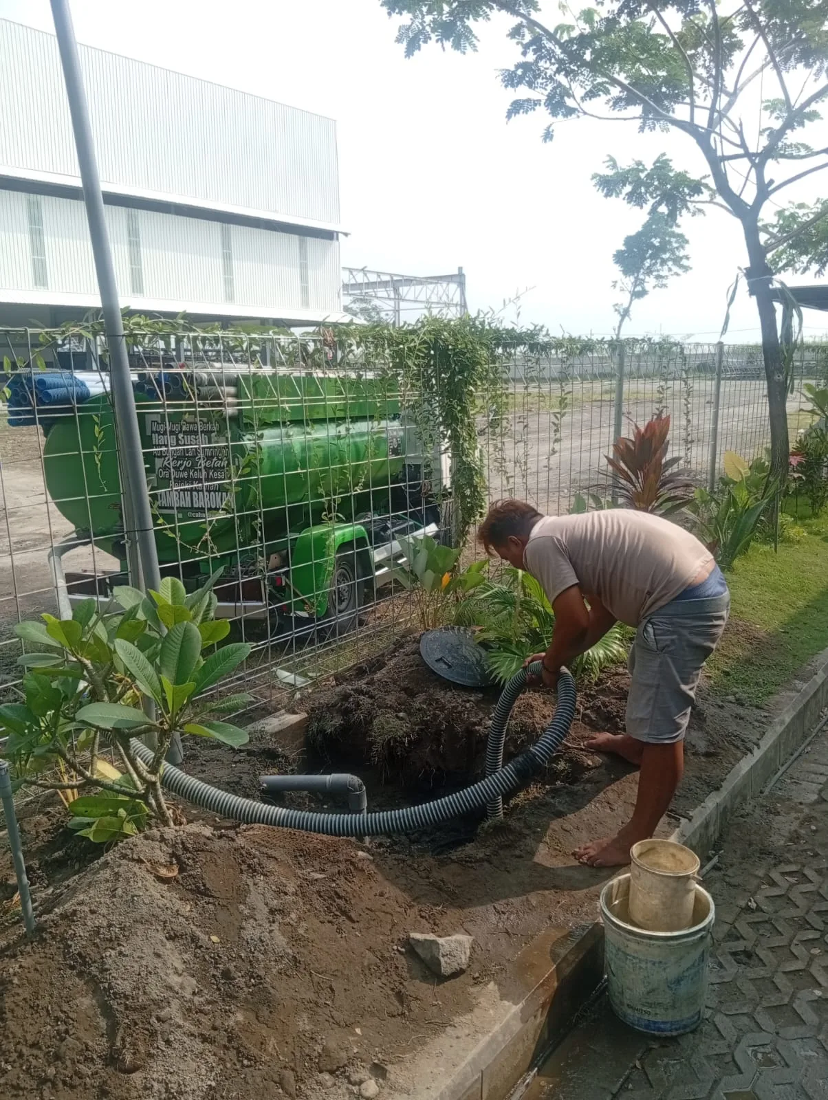 Petugas menangani septic tank di area terbuka dengan truk tangki di belakang.