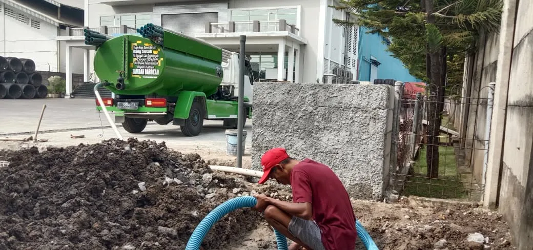 Petugas menyedot septic tank dengan truk tangki hijau di belakang.